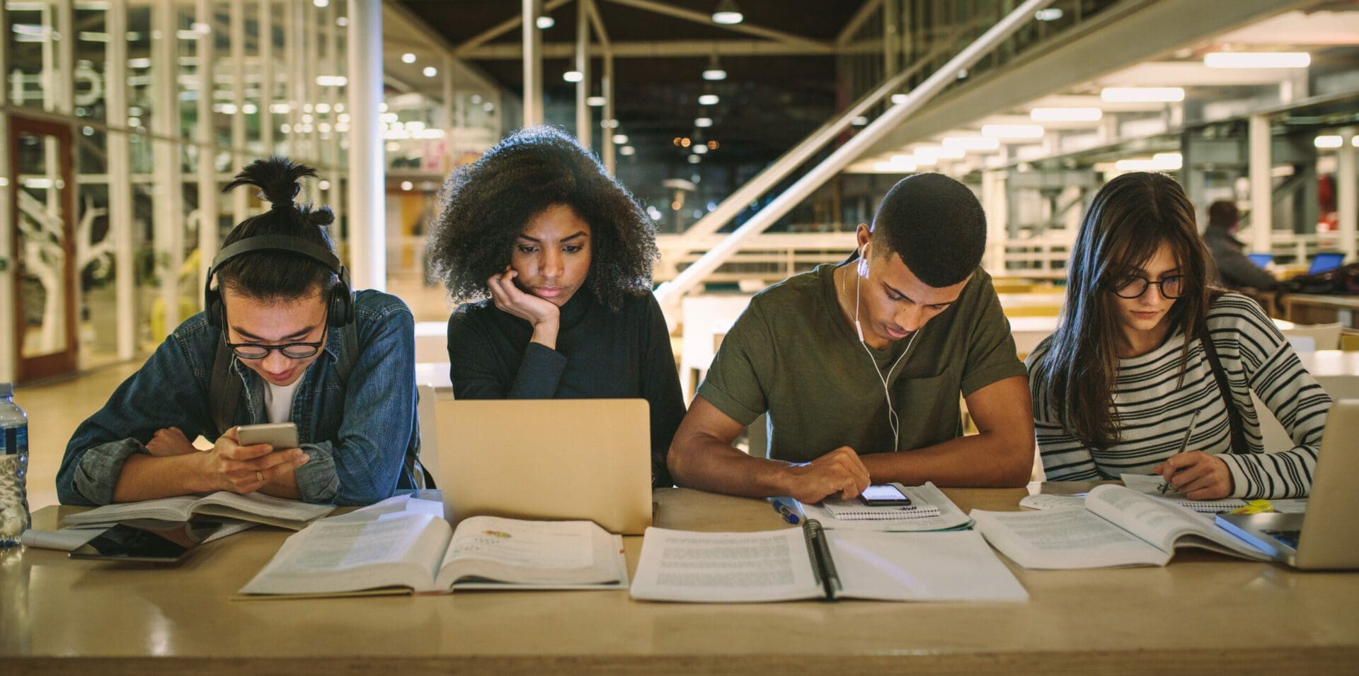 Multi-ethnic students sitting at college library. Students using making notes, using laptop and mobile phones while studying together.