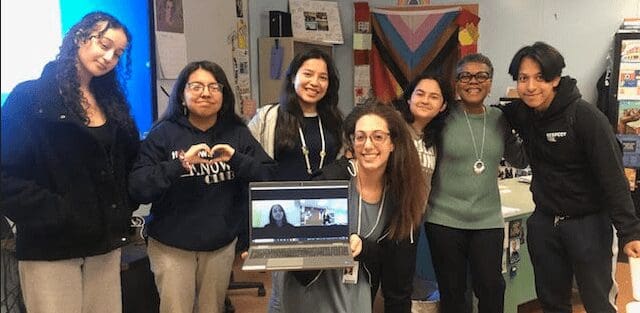 Group of five smiling women posing indoors, one holding a laptop with a video call.