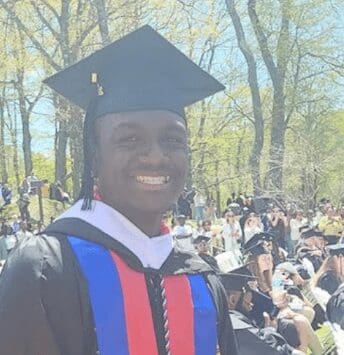 Smiling graduate in cap and gown at outdoor commencement ceremony.