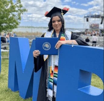 Graduate holding diploma, standing behind large blue letters outdoors.