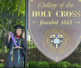 Graduate in cap and gown standing next to Holy Cross College sign.