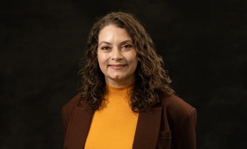 Portrait of a smiling woman with curly hair in a brown blazer and yellow top.