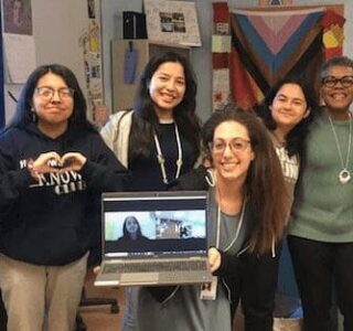 Group of five smiling women posing indoors, one holding a laptop with a video call.