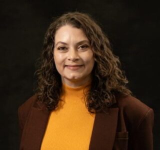 Portrait of a smiling woman with curly hair in a brown blazer and yellow top.